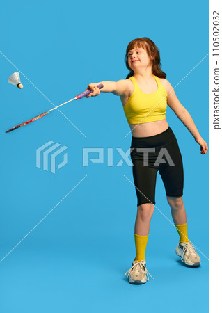 Full-length image of teen girl with down syndrome in sportswear playing badminton, training against blue studio background 110502032