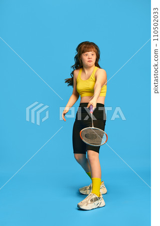 Full-length image of teen girl with down syndrome in sportswear playing badminton, training against blue studio background Full-length image of teen girl with down syndrome in sportswear playing badminton, training against blue studio background 110502033