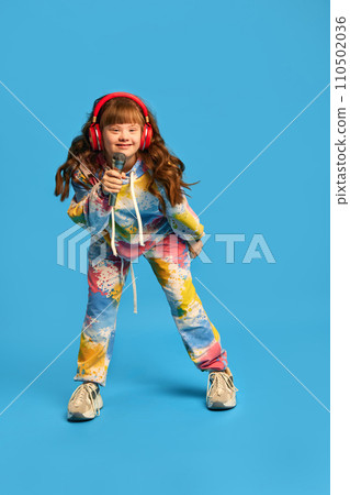 Little happy girl with down syndrome wearing headphones, holing microphone and singing against blue studio background. Hobby Little happy girl with down syndrome wearing headphones, holing microphone and singing against blue studio background. Hobby 110502036