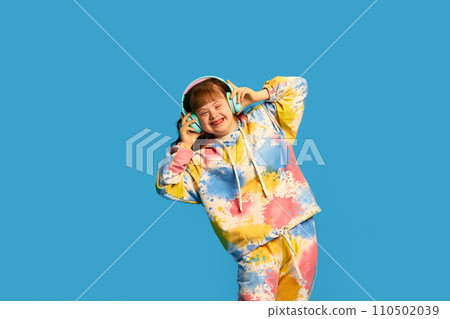 Happy, smiling little girl with down syndrome listening to music in headphones against blue studio background Happy, smiling little girl with down syndrome listening to music in headphones against blue studio background 110502039