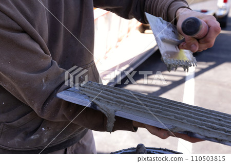 When applying cement adhesive, tiler spreads wet adhesive with trowel on ceramic tile 110503815
