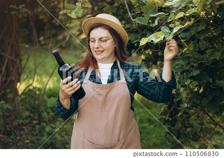 Young happy woman holding a bottle of wine in the grape fields. Ripe grapes in fall. Wine concept Young happy woman holding a bottle of wine in the grape fields. Ripe grapes in fall. Wine concept 110506830