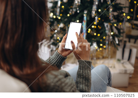 hands holding phone with empty screen on background of golden beautiful christmas tree with lights in evening 110507025