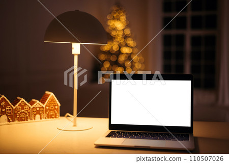 Laptop computer with blank screen on a table in a cozy dark room with Christmas decor. Mockup copy space. Laptop computer with blank screen on a table in a cozy dark room with Christmas decor. Mockup copy space. 110507026
