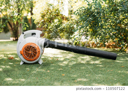A close-up of an orange cordless, electric leaf blower lying on a grass. Autumn, fall gardening works in a backyard, on a lawn. Garden works. 110507122