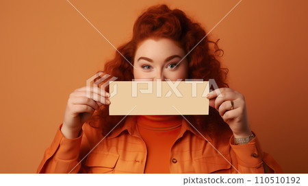 Portrait of beautiful young woman with red curly hair in orange coat holding blank card over her face. Portrait of beautiful young woman with red curly hair in orange coat holding blank card over her face. 110510192