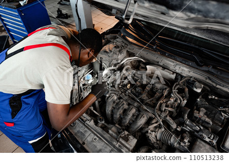 Young African auto mechanic checking car engine under the hood in auto service Young African auto mechanic checking car engine under the hood in auto service 110513238