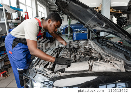 Young African auto mechanic checking car engine under the hood in auto service 110513432