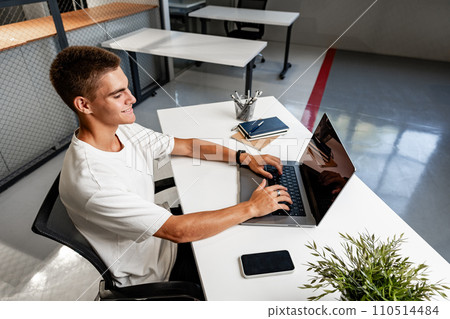Casual young man in white t-shirt sitting at white desk looking at laptop screen at modern office Casual young man in white t-shirt sitting at white desk looking at laptop screen at modern office 110514484