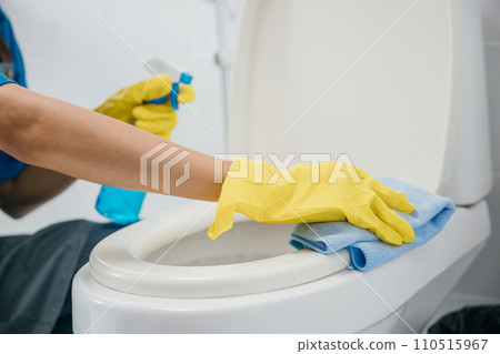 A maid in yellow rubber gloves scrubs toilet seat with cloth ensuring purity and hygiene in the bathroom. Her deep cleaning emphasizes housekeeping and healthcare. A maid in yellow rubber gloves scrubs toilet seat with cloth ensuring purity and hygiene in the bathroom. Her deep cleaning emphasizes housekeeping and healthcare. 110515967