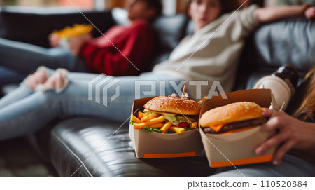 Burgers and fries on the sofa and blurred portrait of teenagers sitting on the sofa and watching TV, fresh food delivery, small local business concept, banner or advertising idea 110520884