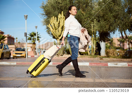 Woman traveler with yellow suitcase, crossing the street on pedestrian crosswalk, confidently looking into the distance. 110523019