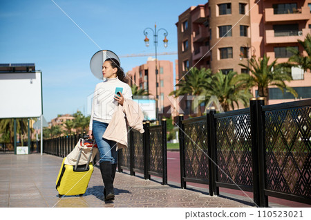 Full length portrait of a woman with yellow luggage, looking aside, holding mobile phone while walking in city street 110523021