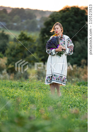 A young girl wearing a long embroidered shirt holds a bouquet of lavender in her hands. A young girl wearing a long embroidered shirt holds a bouquet of lavender in her hands. 110523923