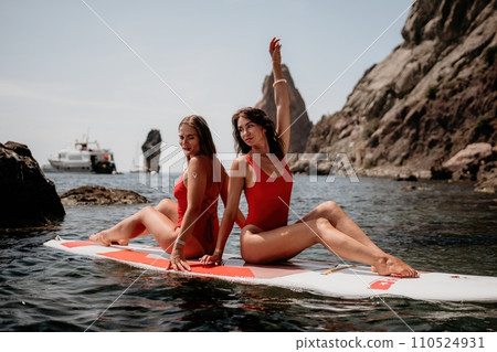 Woman sup yoga. Two happy sporty women practising yoga pilates on paddle sup surfboard. Female stretching doing workout on sea water. Modern individual female hipster outdoor summer sport activity. Woman sup yoga. Two happy sporty women practising yoga pilates on paddle sup surfboard. Female stretching doing workout on sea water. Modern individual female hipster outdoor summer sport activity. 110524931