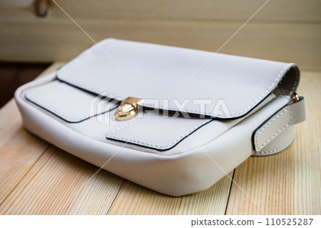 White leather women's handbag with a strap on a light wood background. An elegant accessory for stylish women.. .Handbag made of genuine leather, comfortable and practical. White leather women's handbag with a strap on a light wood background. An elegant accessory for stylish women.. .Handbag made of genuine leather, comfortable and practical. 110525287