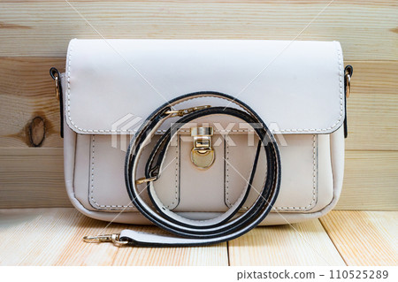 White leather women's handbag with a strap on a light wood background. An elegant accessory for stylish women.. .Handbag made of genuine leather, comfortable and practical. White leather women's handbag with a strap on a light wood background. An elegant accessory for stylish women.. .Handbag made of genuine leather, comfortable and practical. 110525289