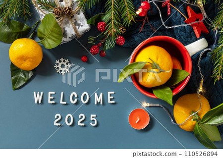 Welcome 2025 text. Winter composition with fir branches, present, cones and tangerines on a blue background. Cozy winter mood. View from above. Welcome 2025 text. Winter composition with fir branches, present, cones and tangerines on a blue background. Cozy winter mood. View from above. 110526894