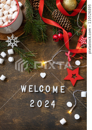 Winter, christmas lights, cozy cup hot chocolate drink with marshmallow on wooden table background. Seasonal relaxing and still life concept. View from above with english text welcome 2024. Winter, christmas lights, cozy cup hot chocolate drink with marshmallow on wooden table background. Seasonal relaxing and still life concept. View from above with english text welcome 2024. 110526895
