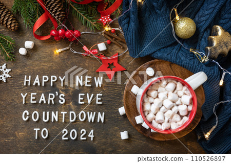 Winter, christmas lights, cozy cup hot chocolate drink with marshmallow on wooden table background. View from above with english text Happy New Year's Eve countdown to 2024. 110526897