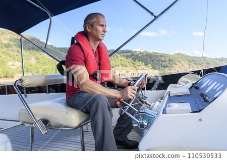 Young caucasian man steering motor yacht. Motor boat captain. Master of yacht. Yachting concept. Man wearing inflatable life jacket. 110530533