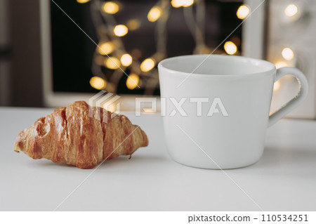 Large croissant on a plate with a large white mug of coffee against the background of a garland, close-up Large croissant on a plate with a large white mug of coffee against the background of a garland, close-up 110534251