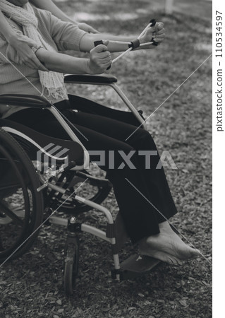 Young nurse or physiotherapist in scrubs helping a happy retired old woman do fitness exercises with light weight dumbbells at home. Concept of physiotherapy for seniors 110534597