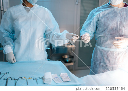 Cropped shot of unrecognizable assistant in surgical uniform assists surgeons, giving medical instruments during surgical operation. Doctors wearing sterile gloves, surgical uniforms and masks 110535464