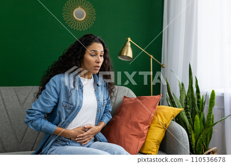 Surprised woman sitting on couch with hand on stomach, expressing shock or discomfort at home with green decor. Surprised woman sitting on couch with hand on stomach, expressing shock or discomfort at home with green decor. 110536678
