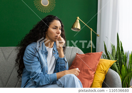 Woman coughing into her hand while sitting on a couch, feeling unwell, in a cozy home environment with indoor plants. 110536680
