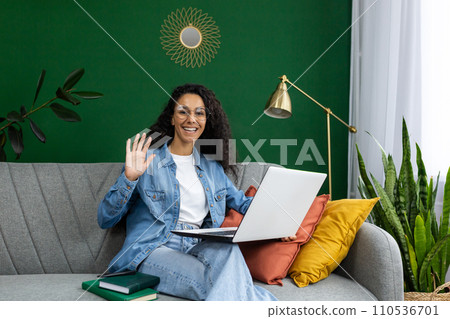 Cheerful woman with glasses waving while holding a laptop, sitting on a couch with colorful cushions and indoor plants. 110536701