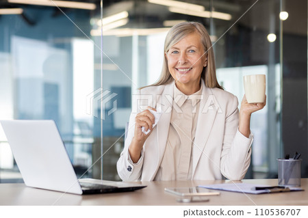 Portrait of a smiling gray-haired businesswoman sitting in the office at the table with a cup and a napkin and smiling at the camera. 110536707