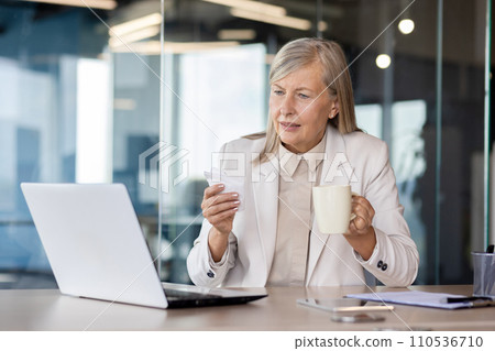 Sick senior business woman working in the office, sitting at the table, holding a cup and a napkin in her hands, looking worriedly at the laptop screen. 110536710