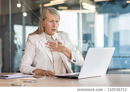 Senior woman in business suit sitting at desk in office in front of laptop and holding hand to chest, having panic attack, heart attack, shocked by business affairs. Senior woman in business suit sitting at desk in office in front of laptop and holding hand to chest, having panic attack, heart attack, shocked by business affairs. 110536712