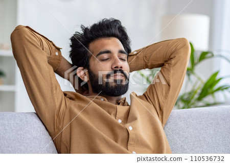 Relaxed bearded man with closed eyes resting on a couch, hands behind head in a cozy room, concept of peace and leisure time at home. Relaxed bearded man with closed eyes resting on a couch, hands behind head in a cozy room, concept of peace and leisure time at home. 110536732