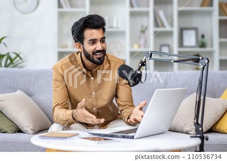 Enthusiastic man podcasting with laptop and microphone at home, expressing positivity and engagement in a cozy setting. Enthusiastic man podcasting with laptop and microphone at home, expressing positivity and engagement in a cozy setting. 110536734