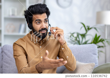 Engaged man with headset in home office conversing lively, hand gesture indicating active communication. Modern remote work lifestyle. 110536742