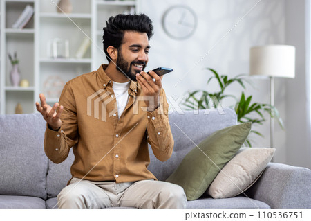 Joyful man speaking into smartphone like a microphone on a cozy sofa, expressing happiness and comfort in a modern living room. 110536751
