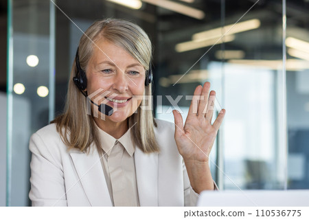 Professional mature woman with headset waving in a friendly manner, representing customer support and positive workplace interaction. Professional mature woman with headset waving in a friendly manner, representing customer support and positive workplace interaction. 110536775