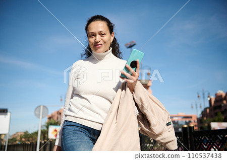 Beautiful woman holding mobile phone while strolling the city street, smiling cutely looking at the camera 110537438