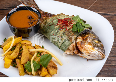 Baked dorado fish in bamboo leaves sprinkled with cilantro and chili threads, baked potatoes and Szechuan sauce. 110538285