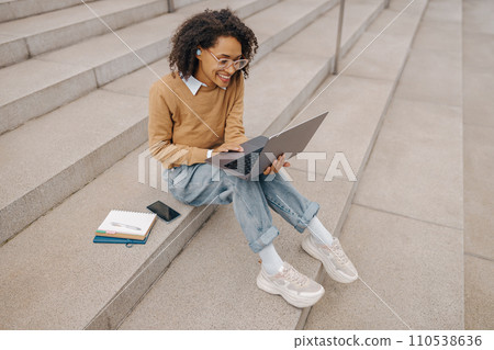 Pretty female entrepreneur working on laptop sitting outside on modern building background  110538636