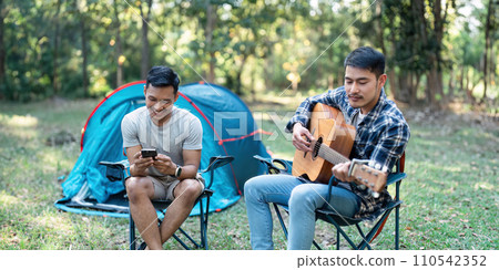 LGBTQIA Gay couple camping together in woods for holidays and relax on guitar together 110542352