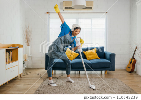 Happy young Asian housewife sings her favorite song with a vacuum cleaner as a microphone while cleaning. Enjoying domestic work dancing in living room a cheerful maid having fun. Happy young Asian housewife sings her favorite song with a vacuum cleaner as a microphone while cleaning. Enjoying domestic work dancing in living room a cheerful maid having fun. 110545239