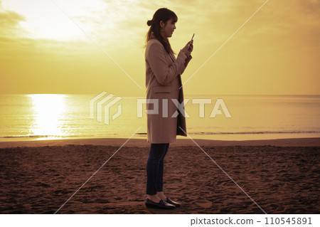 A woman using a smartphone at the winter sea at sunset A woman using a smartphone at the winter sea at sunset 110545891