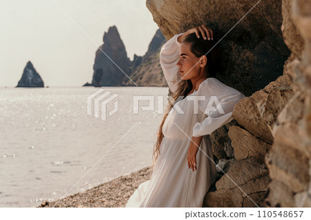 Woman summer travel sea. Happy lady with long hair in white dress enjoy taking photo outdoors for memories. Woman traveler posing on beach at sea surrounded by volcanic mountains, sharing emotions 110548657
