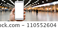 hand holding a mock-up smartphone with a blank screen, set against the dynamic backdrop of an airport interior. This versatile visual is perfect for presenting mobile app designs Generative AI 110552604