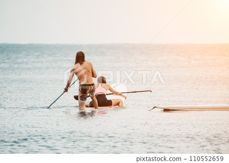 Sea woman and man on sup. Silhouette of happy young woman and man, surfing on SUP board, confident paddling through water surface. Idyllic sunset. Active lifestyle at sea or river. Sea woman and man on sup. Silhouette of happy young woman and man, surfing on SUP board, confident paddling through water surface. Idyllic sunset. Active lifestyle at sea or river. 110552659
