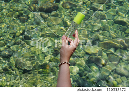 Girl's hand holding cosmetic transparent bottle with body eco spray in front of the sea. fresh marine scented cosmetics mock up 110552872