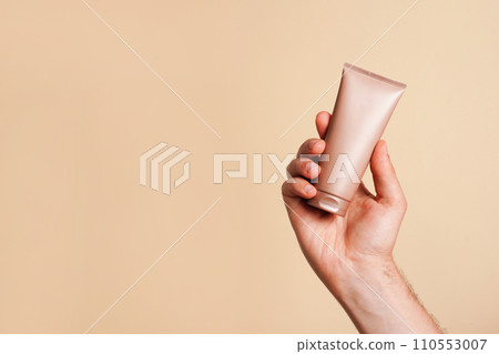 Male hand holding unbranded beige tube with lotion or cosmetics cream on yellow background. Shaving foam or male skin care concept. Minimalism, copy space 110553007